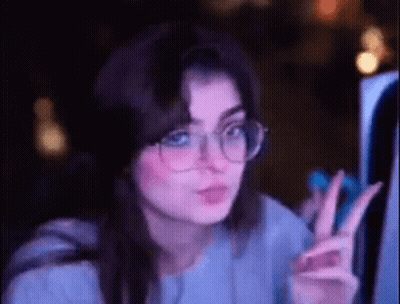 A Woman Wearing Glasses Is Giving A Peace Sign While Looking At A Computer Screen . GIF