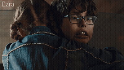 A Woman Wearing Glasses Is Hugging Another Woman Wearing A Blue Shirt With The Name Ezra On The Bottom GIF