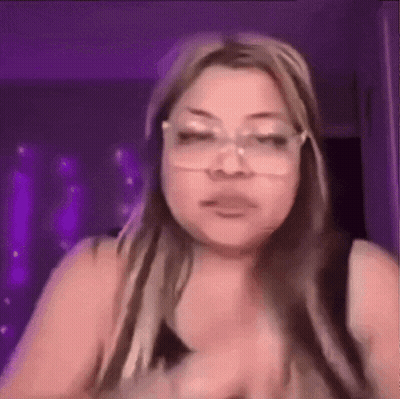 A Woman Wearing Glasses Is Making A Funny Face While Standing In Front Of A Purple Wall . GIF