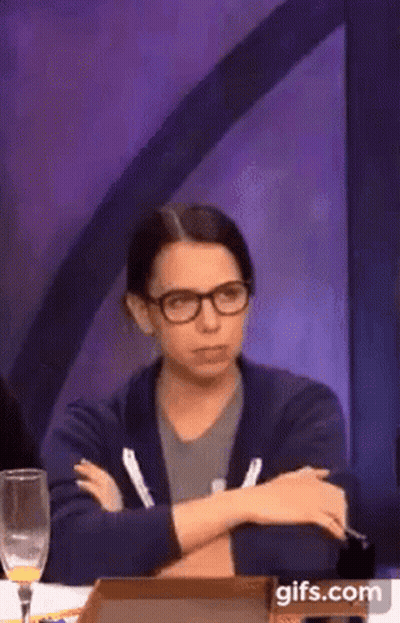 A Woman Wearing Glasses Is Sitting At A Table With Her Arms Crossed And A Glass Of Wine . GIF
