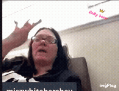 A Woman Wearing Glasses Is Sitting On A Couch And Making A Devil Horns Gesture . GIF
