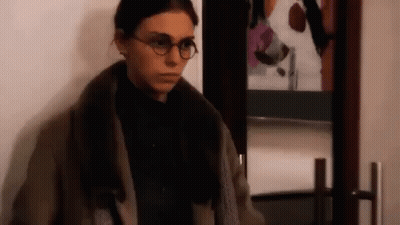 A Woman Wearing Glasses Is Standing In Front Of A Door . GIF