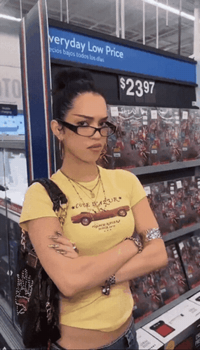 A Woman Wearing Glasses Is Standing In Front Of A Sign That Says Everyday Low Price GIF