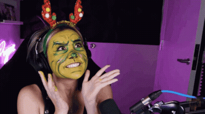 A Woman Wearing Headphones And A Headband With Reindeer Antlers Has A Green And Yellow Face Painted On Her Face GIF