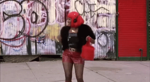 A Woman Wearing Headphones And A Red Head Scarf Stands In Front Of A Graffiti Wall That Says Bo GIF