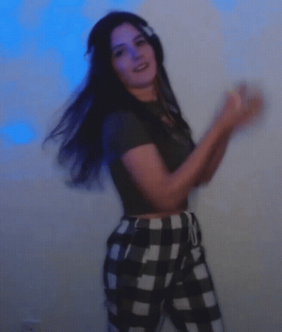 A Woman Wearing Headphones And Plaid Pants Is Dancing In Front Of A Blue Wall . GIF