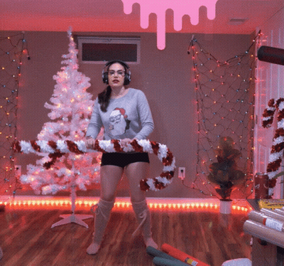 A Woman Wearing Headphones Is Holding A Large Candy Cane In Front Of A Christmas Tree GIF