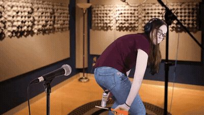 A Woman Wearing Headphones Is Kneeling In Front Of A Microphone And Holding A Can Of Elmer 'S Glue GIF