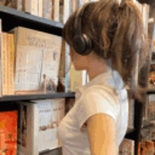 A Woman Wearing Headphones Is Looking At Books In A Library GIF
