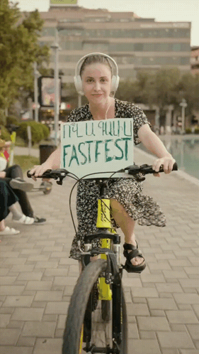 A Woman Wearing Headphones Is Riding A Bike While Holding A Sign That Says Fastfest GIF