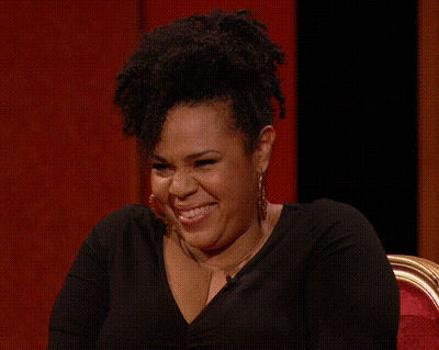 A Woman Wearing Hoop Earrings And A Black Shirt Is Laughing While Sitting In A Chair . GIF