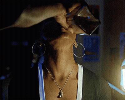 A Woman Wearing Hoop Earrings And A Necklace With A Monkey Pendant Is Drinking From A Glass GIF