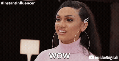 A Woman Wearing Hoop Earrings And A Pink Turtleneck Sweater Is Saying Wow GIF