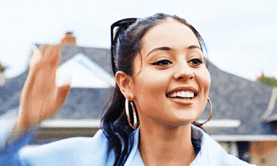 A Woman Wearing Hoop Earrings And A Ponytail Is Smiling In Front Of A House GIF