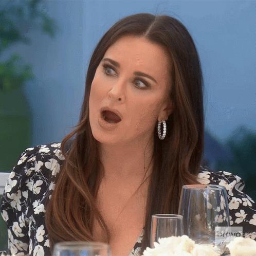 A Woman Wearing Hoop Earrings Is Sitting At A Table With A Bravo Logo In The Background GIF