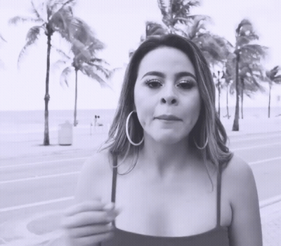 A Woman Wearing Hoop Earrings Is Standing On A Sidewalk In Front Of Palm Trees GIF