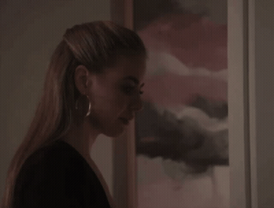 A Woman Wearing Hoop Earrings Stands In Front Of A Framed Picture GIF