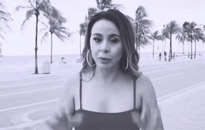 A Woman Wearing Hoop Earrings Stands In Front Of Palm Trees On The Beach GIF