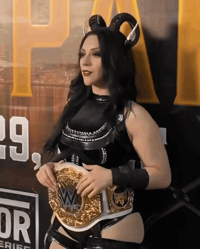 A Woman Wearing Horns And Holding A Wrestling Belt In Front Of A Sign That Says 29 GIF