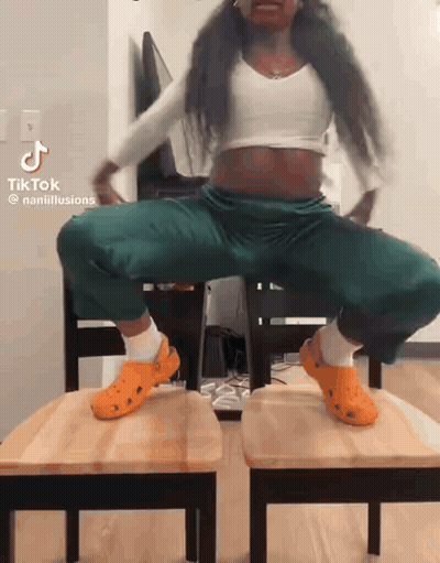 A Woman Wearing Orange Crocs Is Squatting On Two Wooden Chairs .. GIF