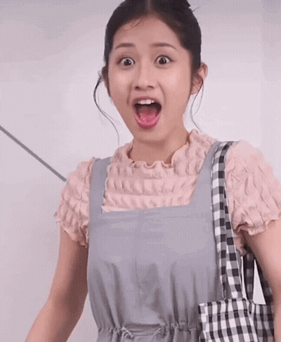 A Woman Wearing Overalls And A Pink Shirt Is Holding A Checkered Bag And Making A Surprised Face GIF
