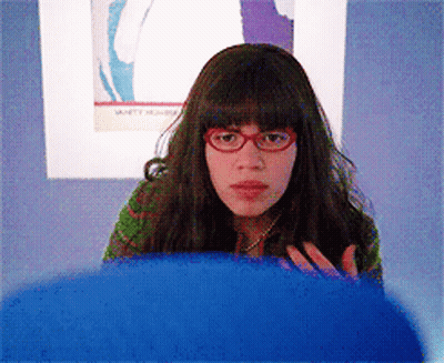 A Woman Wearing Red Glasses Is Sitting In Front Of A Wall With A Picture Of A Woman On It GIF