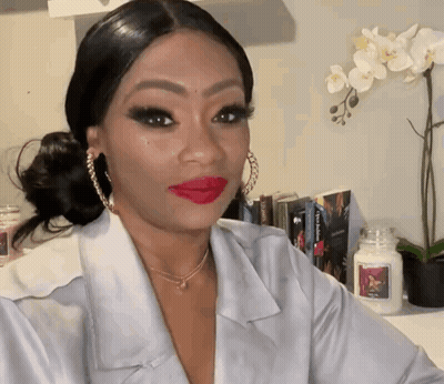 A Woman Wearing Red Lipstick And Hoop Earrings Is Sitting In Front Of A Candle GIF