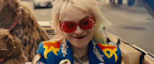 A Woman Wearing Red Sunglasses And A Colorful Jacket Smiles While Sitting In A Car GIF