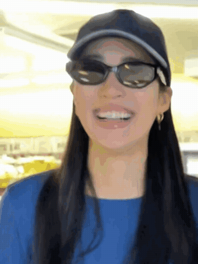 A Woman Wearing Sunglasses And A Baseball Cap Smiles For The Camera GIF