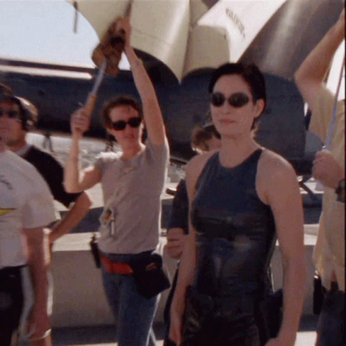 A Woman Wearing Sunglasses And A Black Tank Top Stands In Front Of A Plane That Says " Valiant " On It GIF