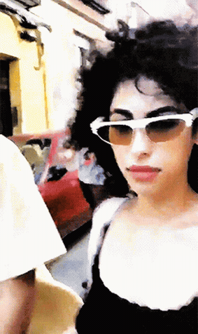 A Woman Wearing Sunglasses And A Black Top Is Walking Down A Street GIF