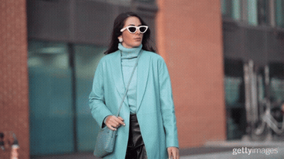 A Woman Wearing Sunglasses And A Blue Coat Is Walking Down The Street GIF