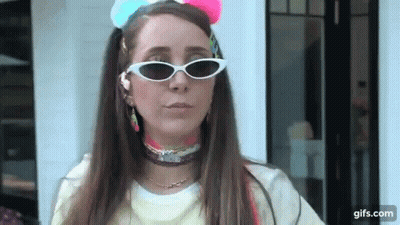 A Woman Wearing Sunglasses And A Colorful Headband Is Standing In Front Of A White Building . GIF