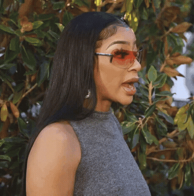 A Woman Wearing Sunglasses And A Gray Top Stands In Front Of A Tree GIF