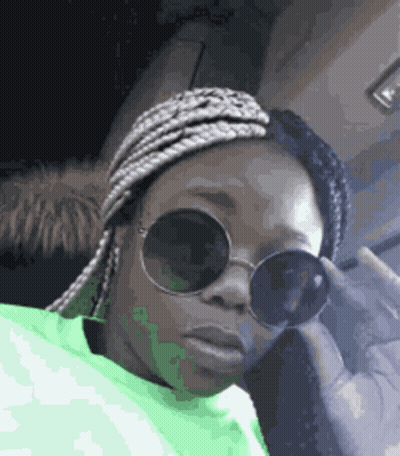 A Woman Wearing Sunglasses And A Green Sweater Is Sitting In A Car . GIF