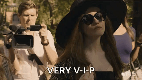 A Woman Wearing Sunglasses And A Hat Is Standing In Front Of A Camera While A Man Takes A Picture . GIF