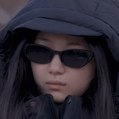 A Woman Wearing Sunglasses And A Hooded Jacket Looks At The Camera GIF