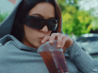 A Woman Wearing Sunglasses And A Hoodie Drinks From A Plastic Cup GIF
