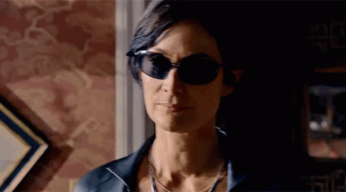 A Woman Wearing Sunglasses And A Necklace Is Standing In A Room . GIF