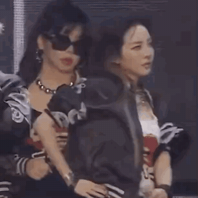 A Woman Wearing Sunglasses And A Necklace Is Standing Next To Another Woman In A Room . GIF
