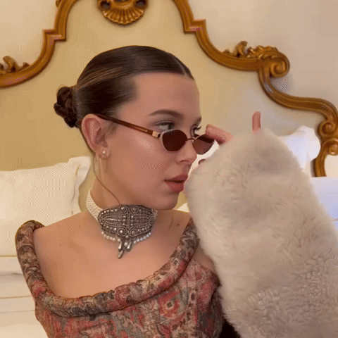 A Woman Wearing Sunglasses And A Pearl Choker Looks Down GIF
