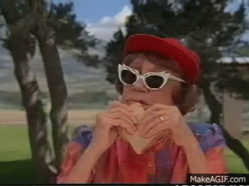 A Woman Wearing Sunglasses And A Red Hat Is Eating A Sandwich On Makeagif.com GIF