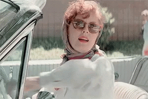 A Woman Wearing Sunglasses And A Scarf Is Driving A White Car . GIF