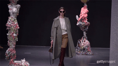 A Woman Wearing Sunglasses And A Trench Coat Walks Down A Runway In Front Of Sculptures GIF