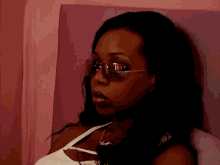 A Woman Wearing Sunglasses And A White Tank Top Is Leaning Against A Pink Wall . GIF