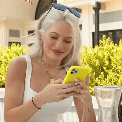 A Woman Wearing Sunglasses And A White Tank Top Is Looking At Her Cell Phone GIF