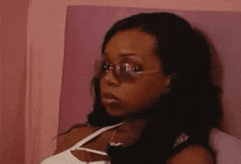 A Woman Wearing Sunglasses And A White Tank Top Is Sitting On A Bed . GIF