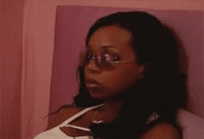 A Woman Wearing Sunglasses And A White Tank Top Is Sitting On A Bed GIF