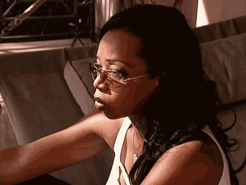 A Woman Wearing Sunglasses And A White Tank Top Is Sitting On A Couch GIF