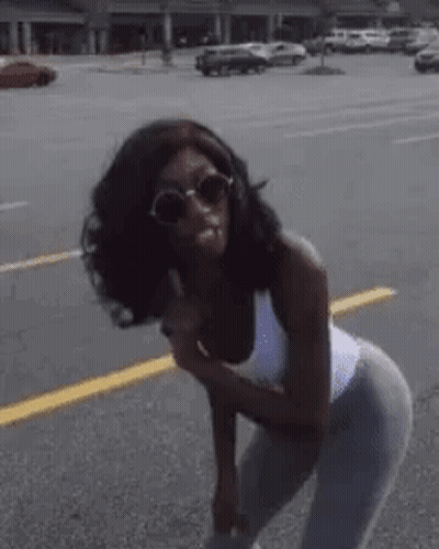 A Woman Wearing Sunglasses And A White Tank Top Is Standing On The Side Of The Road . GIF